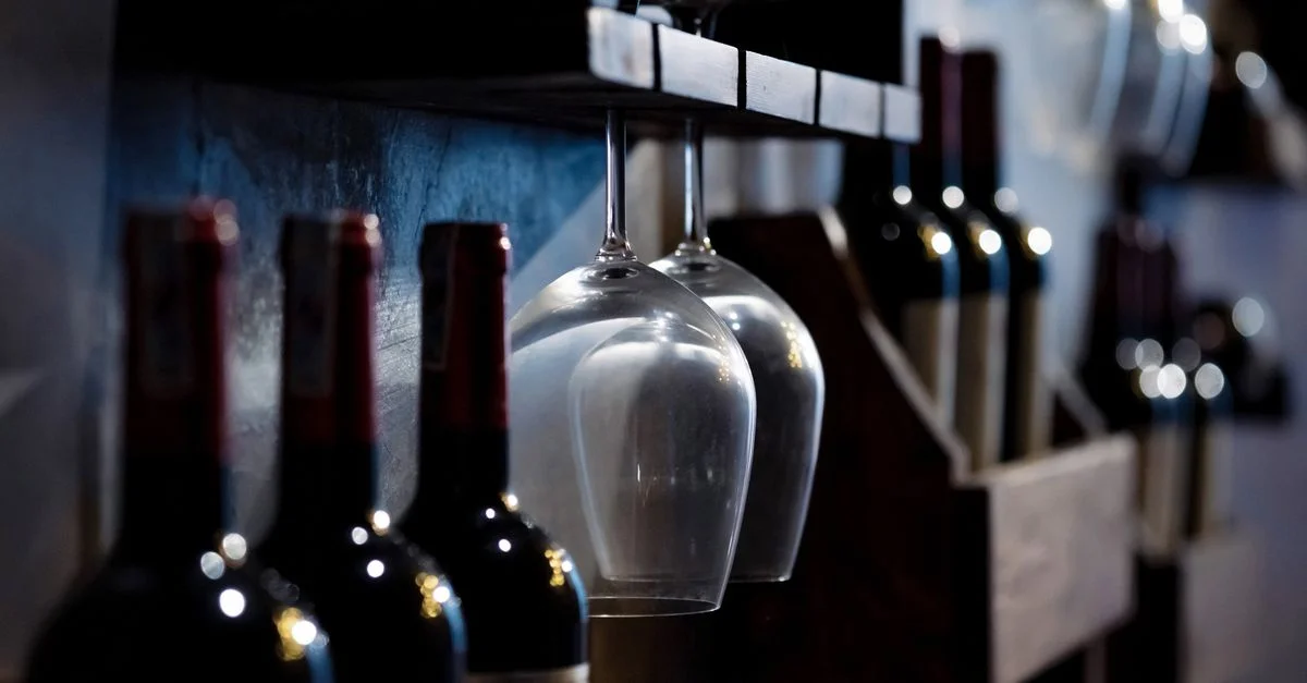 Gros plan de bouteilles et verres de vin sur des étagères en bois dans une cave faiblement éclairée, créant une ambiance chaleureuse.
